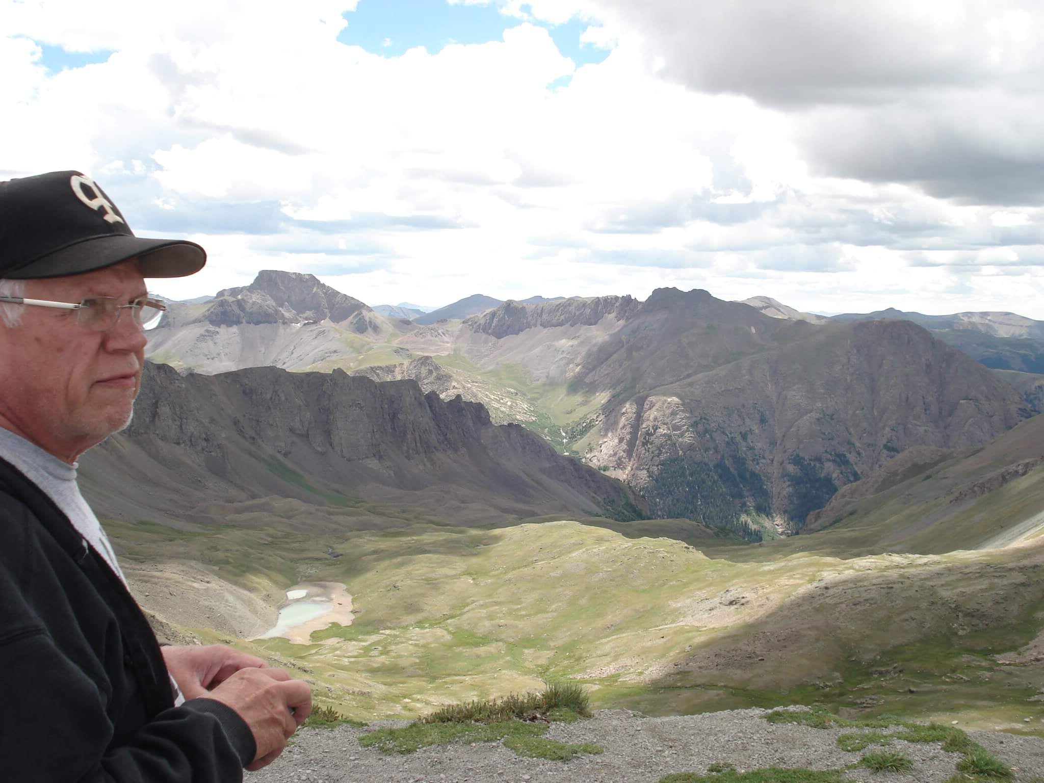 Man wearing a cap and glasses standing in front of a mountainous landscape with clouds.